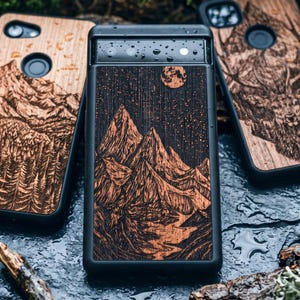 May include: Black phone case with a wood-grain mountain scene. The case features a detailed engraving of a mountain range under a starry night sky with a full moon. The case is surrounded by other cases with similar designs.