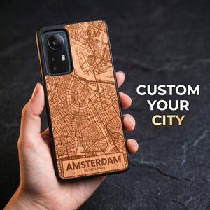 May include: A phone case with a wooden map of Amsterdam, Netherlands, held in a hand. The case is brown with a black border and features the text "CUSTOM YOUR CITY". The city name is printed at the bottom.