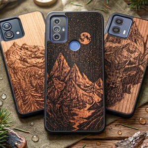 以下が含まれることがあります： 黒い縁取りの木製携帯電話ケース3つ。ケースには、星空の下の山脈、森、鹿の彫刻デザインが施されています。木材は暖かく自然な色合いです。