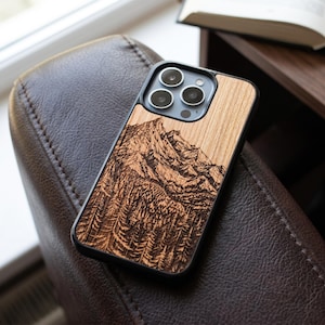 May include: A wooden phone case with a detailed mountain and forest engraving. The case is made of light-colored wood and has a black border. The phone case is resting on a brown leather chair.