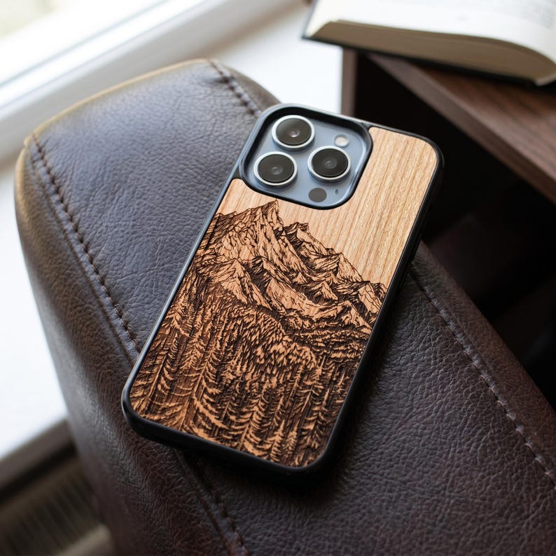May include: A wooden phone case with a detailed mountain and forest engraving. The case is made of light-colored wood and has a black border. The phone case is resting on a brown leather chair.