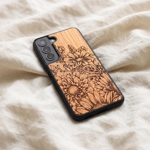 May include: A phone case with a wooden back featuring an engraved floral design. The case has a black border and a camera cutout. The flowers are intricately detailed and set against a light wood grain.