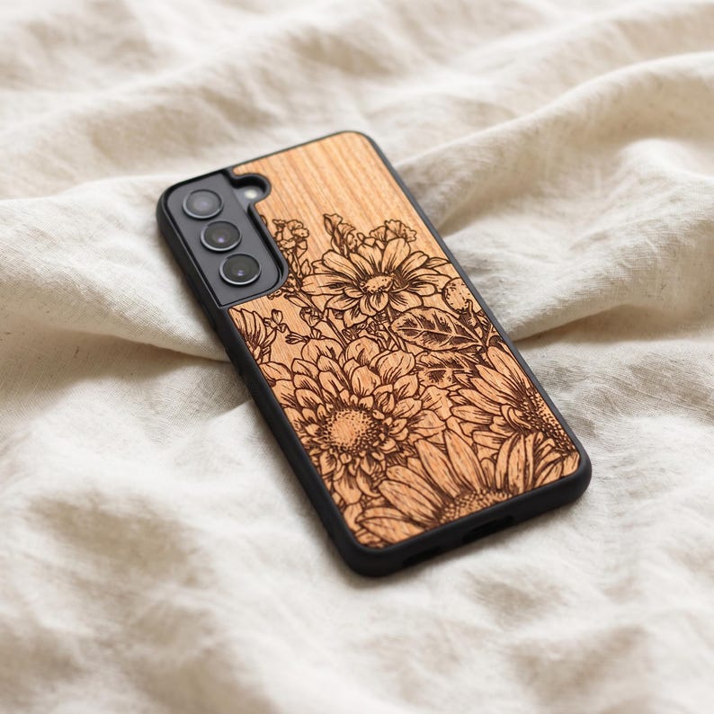 May include: A phone case with a wooden back featuring an engraved floral design. The case has a black border and a camera cutout. The flowers are intricately detailed and set against a light wood grain.