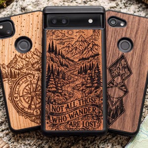May include: Three phone cases with wood-grain back panels featuring laser-engraved nature designs. The center case displays a mountain landscape with a winding river and the text "NOT ALL THOSE WHO WANDER ARE LOST" in a banner at the bottom.
