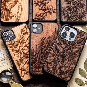 Peut inclure: Une collection de coques pour smartphone avec un dos en bois naturel orné d'illustrations botaniques gravées au laser en noir, représentant des eucalyptus, des fougères, des feuilles de monstera, des magnolias et des branches d'olivier.