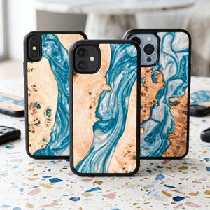 Puede incluir: Varias fundas para teléfono con un diseño único. Tienen un borde negro y un patrón de efecto madera con resina azul y blanca fluida. La veta de la madera es de color marrón claro. Las fundas están dispuestas sobre una superficie moteada.