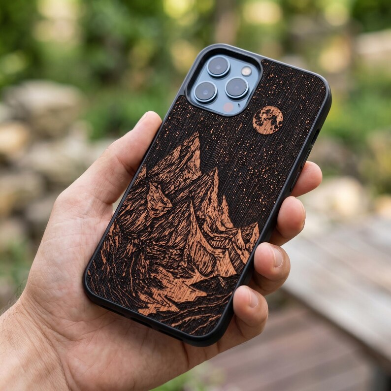 May include: A black phone case with a mountain and starry night design. The case features a detailed wood-grain texture with a full moon and mountain range illustration. The phone case is held in a hand.