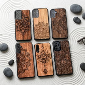 Puede incluir: Seis fundas de teléfono de madera con intrincados diseños de mandala grabados en la madera. Las fundas son de varios tonos marrones y tienen bordes negros. Están dispuestas sobre una superficie blanca texturizada con piedras lisas de color gris oscuro.