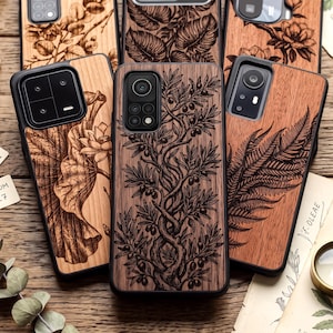 May include: A collection of rectangular phone cases featuring wood-grain back panels with laser-engraved botanical illustrations. The designs include a central olive tree, a lotus flower, and a fern leaf, all encased in black protective frames.