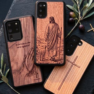Pode incluir: Três capas retangulares para telemóvel com painéis traseiros em grão de madeira. Uma apresenta mãos em oração com um rosário e o texto "If God is for us, who can be against us?". Outra mostra uma figura com um cordeiro. A terceira exibe uma cruz simples.
