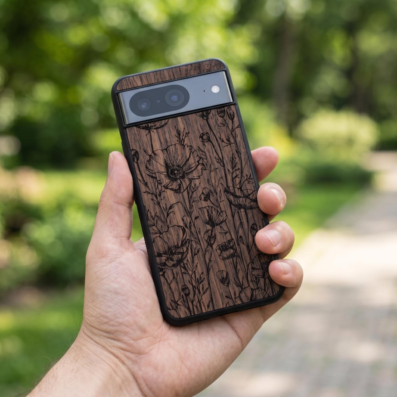 Puede incluir: Funda de tel&eacute;fono de madera marr&oacute;n oscuro con borde negro, con un dise&ntilde;o floral de amapolas. El tel&eacute;fono se sostiene en una mano. La c&aacute;mara del tel&eacute;fono es visible en la parte superior.