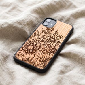 Puede incluir: Funda de tel&eacute;fono de madera con un dise&ntilde;o floral detallado, con girasoles y otras flores. La funda tiene un borde negro y est&aacute; sobre un fondo de tela neutra. La veta de la madera es visible.