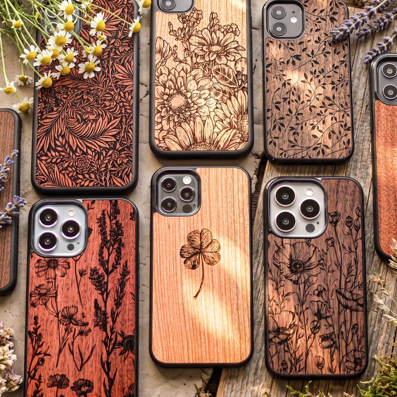 Puede incluir: Una colecci&oacute;n de fundas de tel&eacute;fono de madera con dise&ntilde;os intrincados cortados con l&aacute;ser. Las fundas presentan patrones florales, incluyendo margaritas, hojas y un tr&eacute;bol de cuatro hojas. Cada funda tiene un borde negro y un acabado de veta de madera natural.