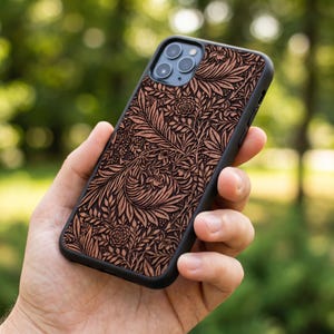 Puede incluir: Una funda de tel&eacute;fono negra con un detallado estampado floral en tonos marrones y beige. La funda, sujeta en una mano, muestra un intrincado dise&ntilde;o de hojas y flores. El tel&eacute;fono tiene tres lentes de c&aacute;mara.