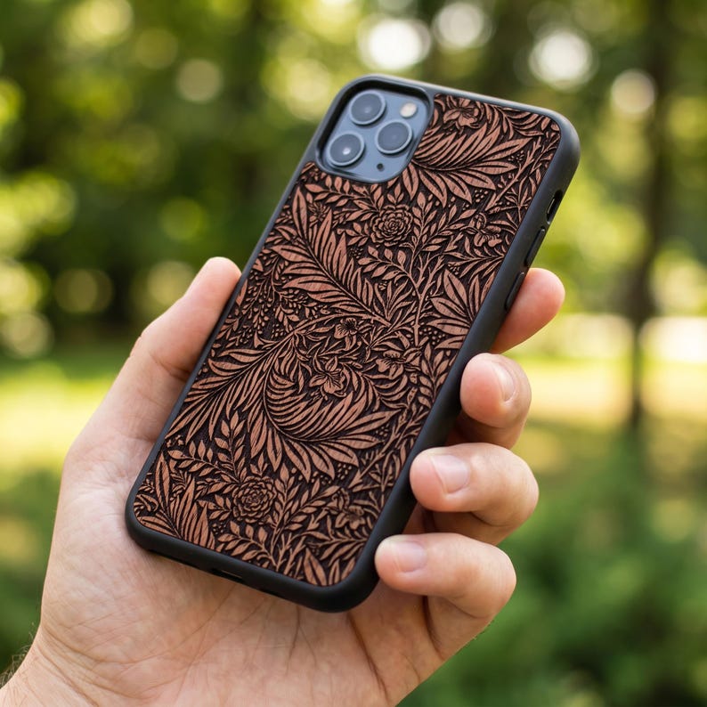 Puede incluir: Una funda de tel&eacute;fono negra con un detallado estampado floral en tonos marrones y beige. La funda, sujeta en una mano, muestra un intrincado dise&ntilde;o de hojas y flores. El tel&eacute;fono tiene tres lentes de c&aacute;mara.