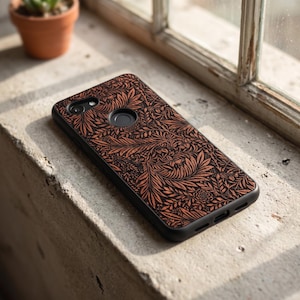 Puede incluir: Funda de tel&eacute;fono inteligente negra con un patr&oacute;n floral detallado en tonos marr&oacute;n y naranja. La funda est&aacute; sobre una superficie de hormig&oacute;n cerca de una ventana. El dise&ntilde;o presenta hojas y flores.