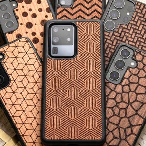 Op de afbeelding: Een collectie smartphonehoesjes met diverse houtnerftexturen en geometrische patronen. Elk hoesje heeft een zwart frame en een achterpaneel met lasergegraveerde ontwerpen, waaronder cirkels, zigzags, kubussen en onregelmatige, steenachtige vormen.