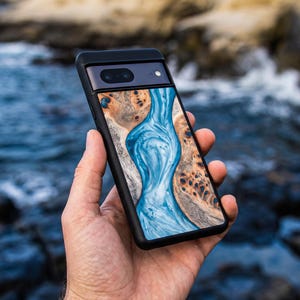 May include: A smartphone case featuring a central inlay of swirling blue resin flanked by natural light brown wood burl with dark knots. The case has a black frame and a horizontal camera bar across the top section of the device back.