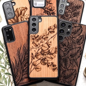 May include: A collection of five smartphone cases featuring natural wood back panels with intricate black laser-engraved botanical illustrations, including ferns, eucalyptus branches, monstera leaves, and floral patterns, all encased in black frames.