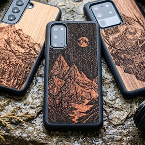 Könnte beinhalten: Handyhüllen aus Holz mit lasergravierten Designs. Eine Hülle zeigt eine Bergkette unter einem Sternenhimmel mit Vollmond. Andere zeigen Berg- und Waldszenen. Die Hüllen haben schwarze Ränder und werden auf einer strukturierten, steinartigen Oberfläche präsentiert.