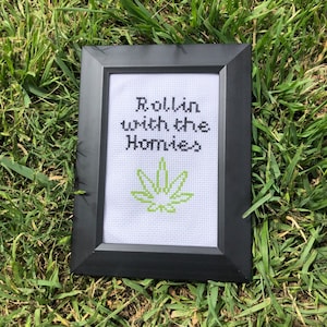 May include: A framed cross-stitch with a green marijuana leaf and the text "Rollin with the Homies".