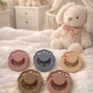 May include: A set of five silicone pacifiers arranged in a semi-circle. The pacifiers feature a round nipple, a circular shield with a scalloped edge, and a central handle. The colors include dusty rose, slate blue, beige, tan, and terracotta.