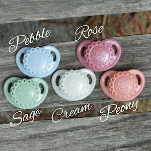 以下が含まれることがあります： 5色の異なる色の哺乳瓶: セージグリーン、クリーム、ローズ、ペブルブルー、ピーチ。各哺乳瓶は、中央に小さな隆起した画像を持つ、テクスチャードの円形デザインです。