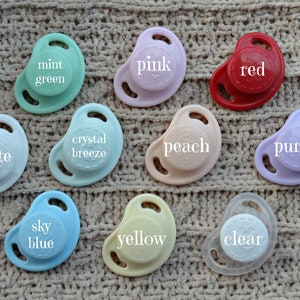 Op de afbeelding: Tien fopspeen in verschillende kleuren, waaronder wit, mintgroen, kristallen bries, perzik, paars, rood, roze, hemelsblauw, geel en helder. Elke fopspeen heeft een andere kleur en een ander tekstlabel.