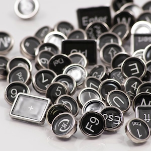 May include: A collection of vintage typewriter keys in black and silver, featuring letters, numbers, and symbols. The keys are scattered on a white surface.