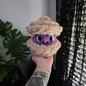 May include: A plush toy shaped like a clam or oyster, featuring two textured, beige, shell-like halves made of soft yarn. Between the shells is a purple, textured center with a speckled appearance, two large black eyes, and a small black stitched smile.