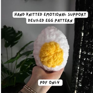 May include: A hand-knitted devilled egg pattern in white and yellow. The egg features a fluffy white exterior and a bright yellow yolk. Text on the image reads "HAND KNITTED EMOTIONAL SUPPORT DEVILED EGG PATTERN" and "PDF ONLY".