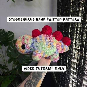 May include: A plush Stegosaurus toy made of multi-colored yarn with red dorsal plates and black eyes. The text STEGOSAURUS HAND KNITTED PATTERN and VIDEO TUTORIAL ONLY is displayed on the image. The toy features a soft, fuzzy texture throughout.