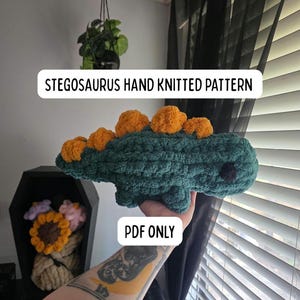 May include: A teal green hand-knitted stegosaurus with orange plates. The text "STEGOSAURUS HAND KNITTED PATTERN" is displayed above the dinosaur. The text "PDF ONLY" is displayed below the dinosaur.