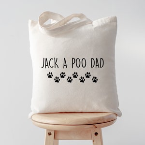 Puede incluir: Bolso tote de color crema con el texto "JACK A POO DAD" en negro, junto con una fila de huellas de patas negras. El bolso tiene un diseño sencillo y un asa para llevar.