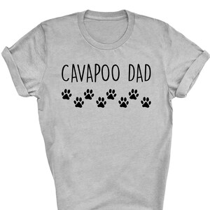 Op de afbeelding: Een grijs T-shirt met de tekst "CAVApoo DAD" en een rij poot afdrukken.