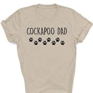 May include: A beige t-shirt with the text "Cockapoo Dad" and a row of black paw prints.