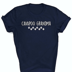 Op de afbeelding: Een marineblauw t-shirt met de tekst "CAVAPOO GRANDMA" in witte letters en een rij witte poot afdrukken onder de tekst.