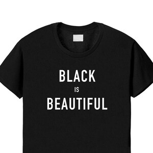 Puede incluir: Una camiseta negra de manga corta con el texto blanco "BLACK IS BEAUTIFUL" impreso en una fuente sans-serif en negrita en el centro del pecho. La prenda tiene un cuello redondo cl&aacute;sico y un corte est&aacute;ndar sobre un fondo liso.