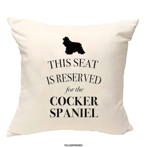 Può includere: Un cuscino bianco con testo nero che recita "Questo posto è riservato al Cocker Spaniel". Sopra il testo c'è una silhouette nera di un Cocker Spaniel.