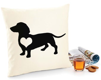 Puede incluir: Una almohada blanca con una silueta negra de un dachshund con un corazón blanco en el pecho. La almohada está sobre una superficie blanca con una taza de té y galletas.