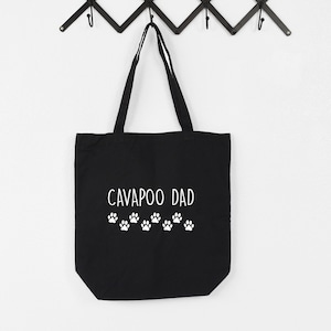 Op de afbeelding: Zwarte canvas tas met witte tekst "Cavapoo Dad" en een rij witte poot afdrukken.