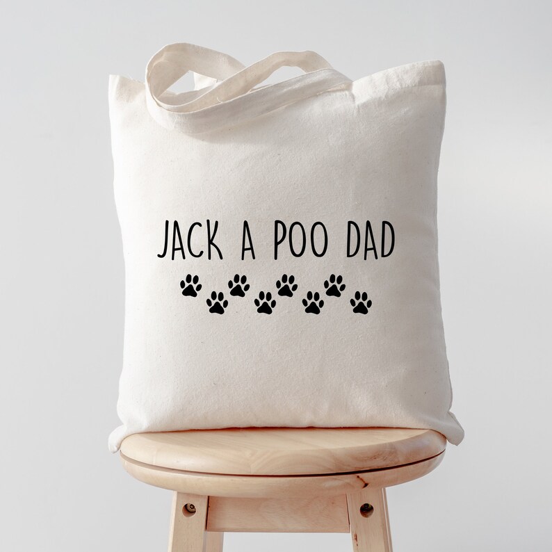 Pu&ograve; includere: Borsa tote color crema con la scritta "JACK A POO DAD" in nero, insieme a una fila di impronte di zampe nere. La borsa ha un design semplice e un manico per il trasporto.