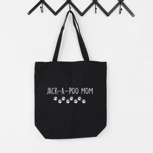Puede incluir: Bolsa de lona negra con texto blanco que dice "JACK-A-POO MOM" y una fila de huellas de patas blancas debajo del texto.