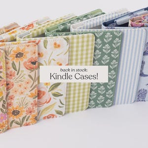 May include: A selection of Kindle cases with diverse floral and patterned designs. The cases showcase a variety of colours, including pink, yellow, green, and blue. The text "back in stock: Kindle Cases!" is displayed on a white label.
