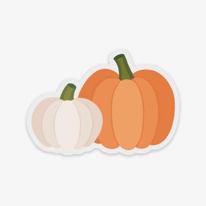 May include: A sticker featuring two pumpkins. One is a light orange color, and the other is white. Both pumpkins have green stems. The sticker has a white border and is set against a white background. The pumpkins are a fall or autumn theme.