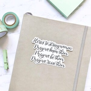May include: A white sticker with black calligraphy that reads "Here's to strong women. May we know them. May we be them. May we raise them."