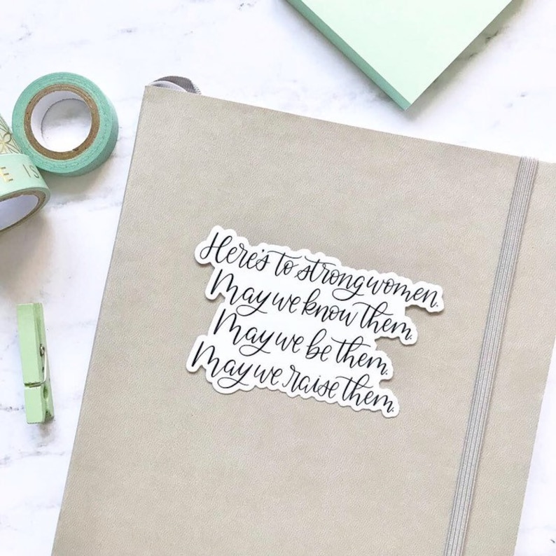 May include: A white sticker with black calligraphy that reads "Here's to strong women. May we know them. May we be them. May we raise them."