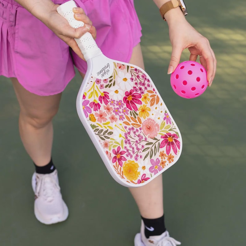 May include: A white pickleball paddle with a floral design in pink, yellow, and orange. The paddle is held by a person, along with a pink pickleball. The text "elyse breanne" is printed on the paddle.