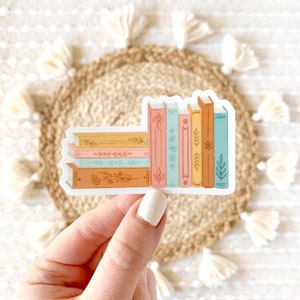 May include: A sticker of a stack of colorful books with floral designs. The books are arranged vertically, with the tallest book on the left and the shortest book on the right. The books are all different colors, including pink, blue, yellow, and brown. The books have floral designs on their covers, including leaves, flowers, and vines.