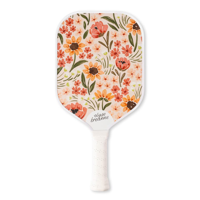 May include: A white pickleball paddle with a floral design featuring sunflowers and other flowers in shades of orange, red, and peach. The paddle has a white handle with a textured grip, and the brand name "elyse breanne" is printed on the paddle.